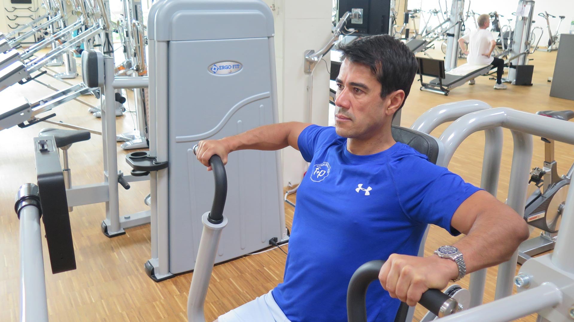 Trainer macht an einem Trainingsgerät eine die Rückenmuskulatur stärkende Übung vor