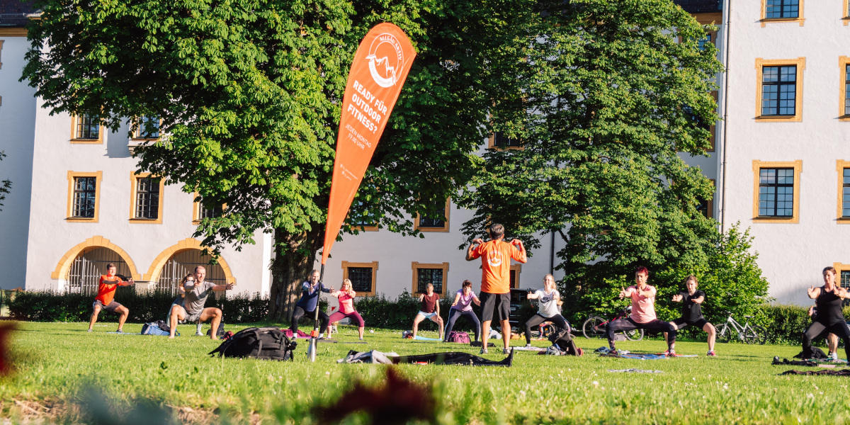 Menschen absolvieren gemeinsam ein funktionelles Ganzkörpertraining auf einer Wiese vor einem historischen Gebäude.